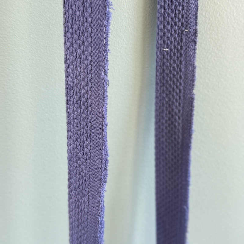 A close-up of two narrow purple woven straps or cords hanging vertically against a pale mint-green background.