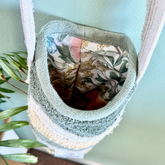 A close-up of a fabric hanging pouch or bag with a soft green terry cloth rim, cream woven trim, and a floral lining, shown against a pale blue wall with plant leaves at the side.