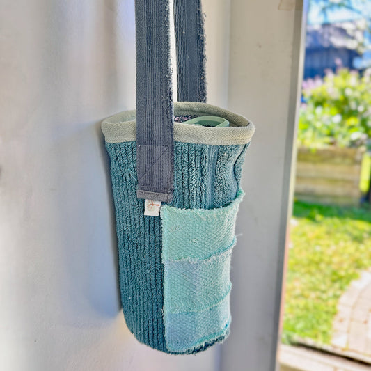 A teal and light blue textured fabric tote bag with a gray strap, hanging on a white door with a garden visible in the background.