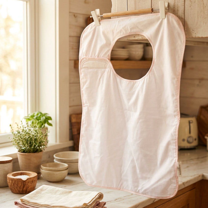A light pink baby bib with a circular neck opening, hanging from a wooden hanger with white clips against an off-white wall.