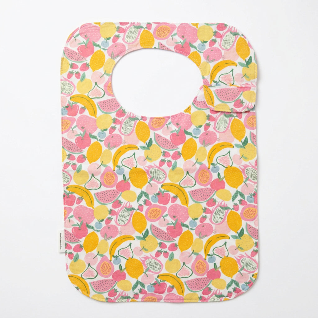 A fabric adult bib with a colourful fruit print, hanging from white clips on a wooden hanger against a light wall.