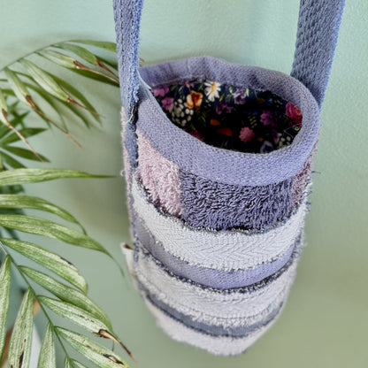 A handmade fabric tote bag with horizontal stripes in lavender, light gray, and pink, featuring a floral lining and a matching woven strap, hanging against a mint green wall next to a green plant.