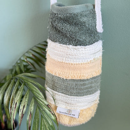 A striped bath towel bag with white shoulder straps, featuring horizontal stripes of sage green, cream, pale yellow, and sage green. It hangs against a sage green wall next to a green palm plant.