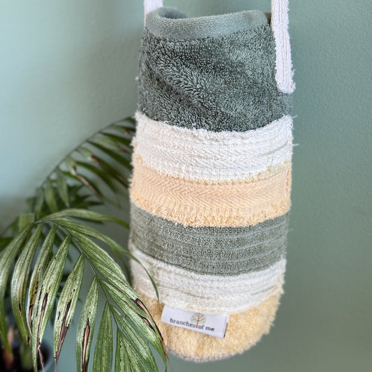 A striped bath towel bag with white shoulder straps, featuring horizontal stripes of sage green, cream, pale yellow, and sage green. It hangs against a sage green wall next to a green palm plant.