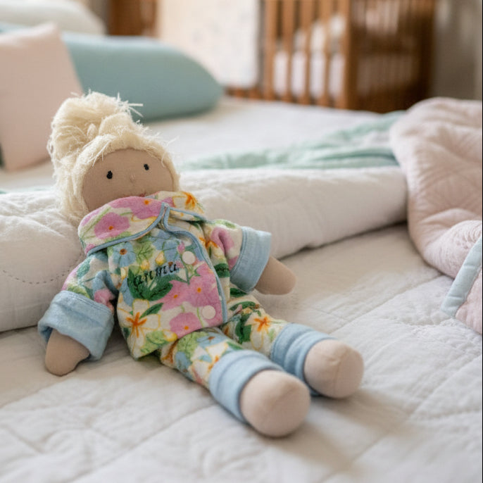A handmade plush doll with messy blonde hair, wearing a colorful floral pajama set with light blue cuffs and a bow, lying on a gray fabric background.