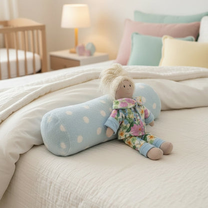 A plush doll with blonde hair lies on a light blue polka-dot bolster pillow on a dark gray bed, wearing a floral pajama set.