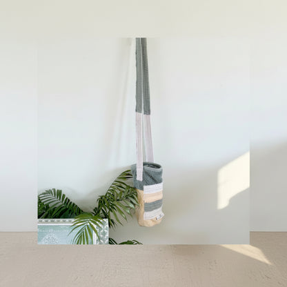 A handmade woven shoulder bag with a long gray and white striped strap, hanging against a light blue wall next to green plant leaves.