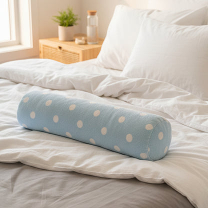 A light blue cylindrical bolster pillow with white polka dots, held by a hand on a dark gray fabric background.