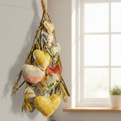 A hanging decorative garland made of multiple fabric hearts in different prints and colors, attached to a natural twig branch and suspended by a rope against a plain white wall.
