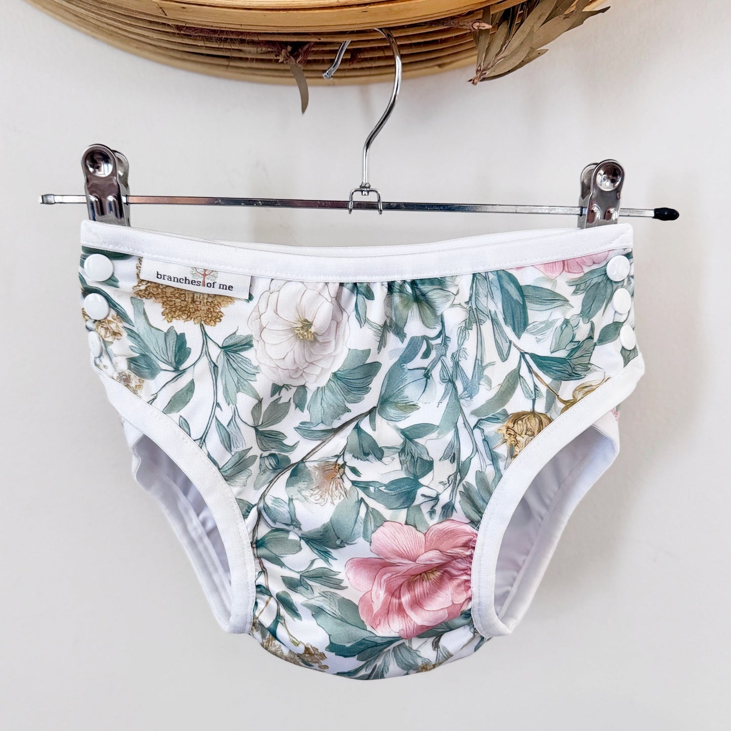 A pair of reusable cloth menstrual pads with a floral print, hanging from a metal hanger on a white wall.
