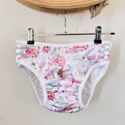 A pair of white swim briefs with a colorful mermaid and sea-themed print, hanging from a metal hanger with black clips against a light beige wall.