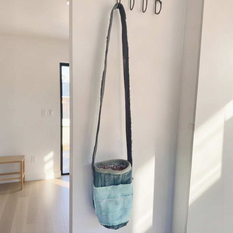 A small crossbody bag with a dark strap and a body made from layered denim in dark blue and light aqua, hanging from a black wall hook on a white wall.