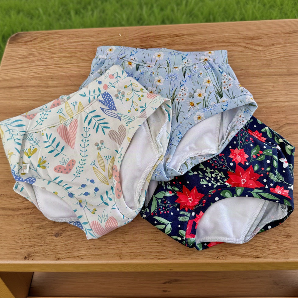 Three reusable cloth swim diapers or brief-style swimwear pieces laid flat on a wooden surface. One has a white background with pastel floral and heart motifs, one has a light blue background with small white flowers and blue accents, and one has a dark navy background with large red floral prints and green leaves.