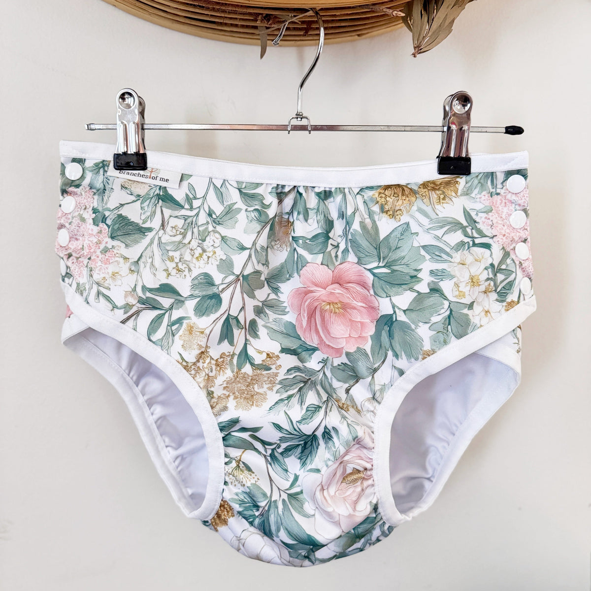 Floral print high-waisted briefs with a white waistband and leg openings, hanging from a metal hanger with black clips against a light wall.