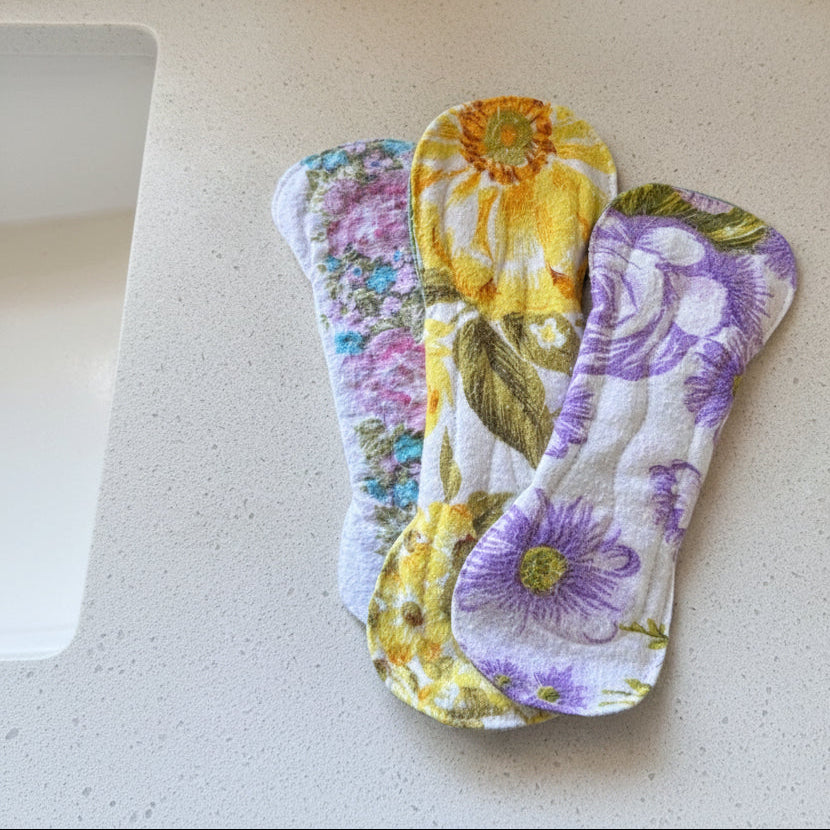 Three reusable cloth menstrual pads with floral prints are arranged on a light speckled countertop. The top pad has a yellow and green floral pattern, while the middle and bottom pads feature yellow sunflowers and purple flowers with green leaves on a white background.