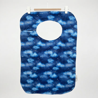 A reusable adult bib with a blue night-sky pattern featuring clouds and stars, hanging from a wooden hanger with white clips against a plain wall.
